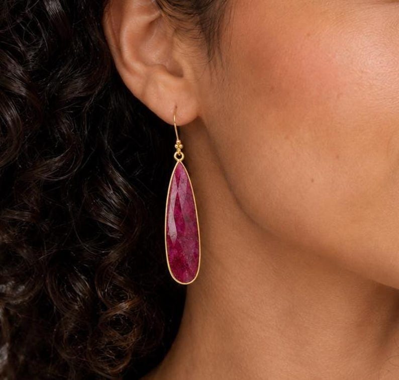 May include: A teardrop-shaped earring featuring a faceted, deep magenta-pink gemstone set in a thin gold-toned metal frame. The earring hangs from a delicate gold-toned hook, showcasing a textured surface on the stone that catches the light.