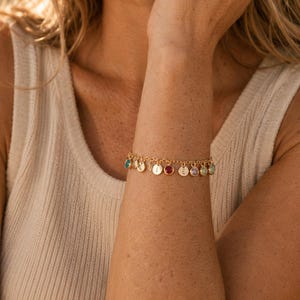 May include: A gold-toned chain bracelet featuring a series of small, dangling charms. The charms alternate between circular metal discs engraved with letters and round, bezel-set faceted gemstones in various colors including red, blue, and green.