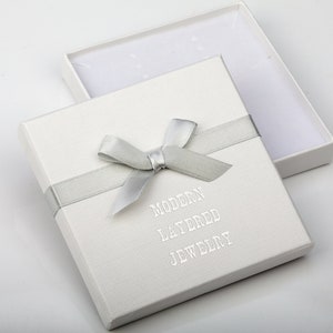 May include: A square white jewelry gift box with a textured surface, featuring a silver ribbon tied in a bow across the top. The text MODERN LAYERED JEWELRY is printed in silver on the lid. The box is open, revealing a white interior lining.