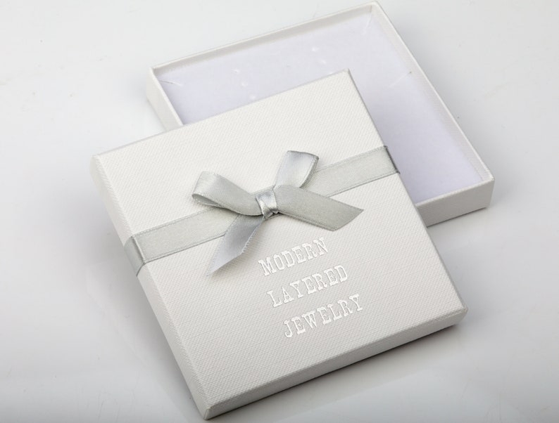 May include: A square white jewelry gift box with a textured surface, featuring a silver ribbon tied in a bow across the top. The text MODERN LAYERED JEWELRY is printed in silver on the lid. The box is open, revealing a white interior lining.
