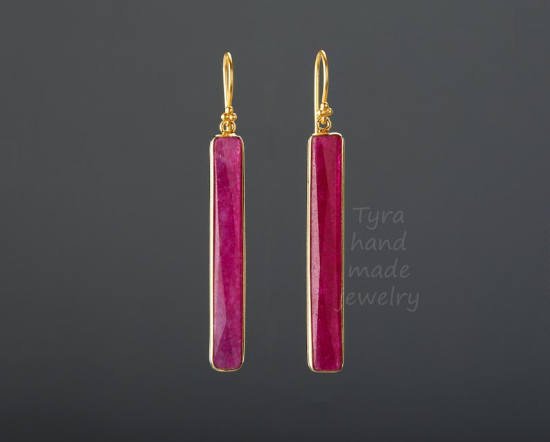 May include: A pair of gold earrings with long, rectangular, pink gemstone drops. The earrings are hanging from a grey background.