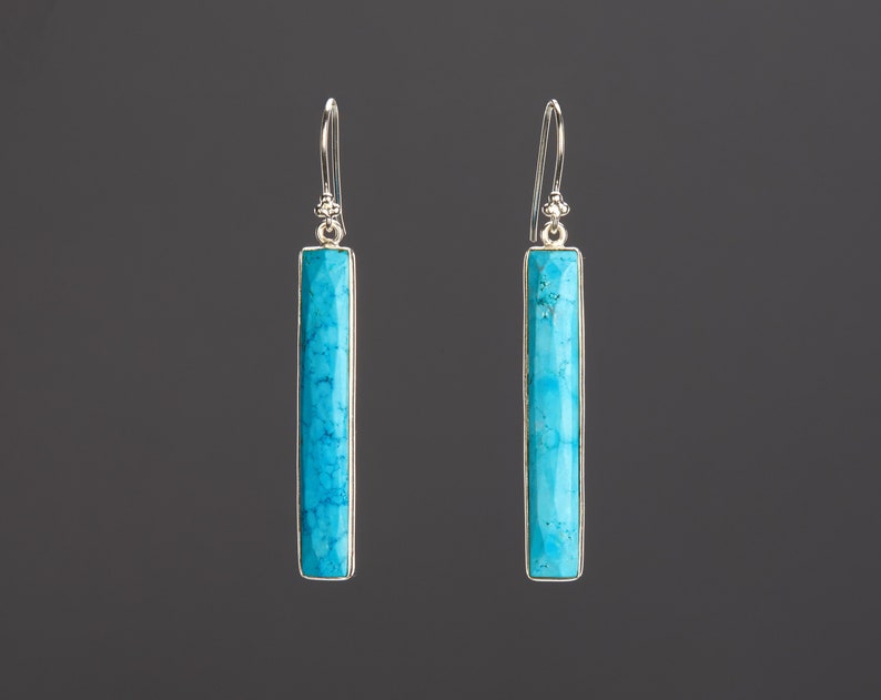 May include: A pair of drop earrings featuring long, rectangular turquoise stones with natural veining. Each stone is set in a polished silver-toned metal frame and hangs from a simple silver-toned hook wire with a small decorative bead detail at the top.