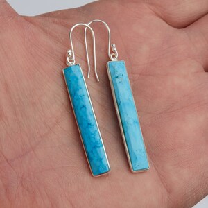 May include: A pair of long, rectangular drop earrings featuring bright blue turquoise stones set in polished silver frames. Each earring hangs from a simple silver hook wire, showcasing a smooth, faceted surface with natural dark veining.