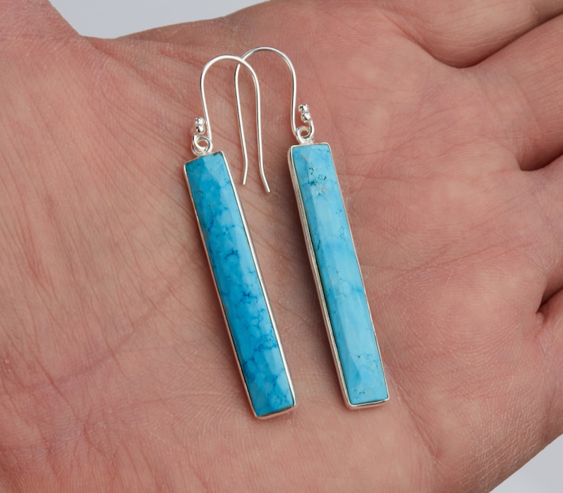 May include: A pair of long, rectangular drop earrings featuring bright blue turquoise stones set in polished silver frames. Each earring hangs from a simple silver hook wire, showcasing a smooth, faceted surface with natural dark veining.