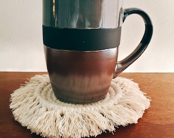 May include: A brown and black ceramic mug with a black band around the middle sits on a white crocheted coaster with fringe.