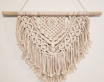 May include: A beige macrame wall hanging with a wooden dowel at the top and a fringe border. The macrame is in a triangular shape with a geometric pattern.