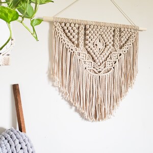May include: A beige macrame wall hanging with a geometric pattern and fringe. The hanging is attached to a wooden dowel rod.