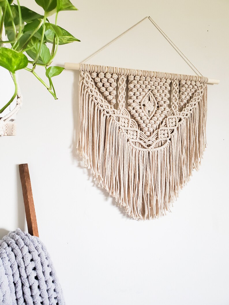 May include: A beige macrame wall hanging with a geometric pattern and fringe. The hanging is attached to a wooden dowel rod.