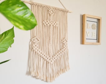 May include: A beige macrame wall hanging featuring an anchor design. The nautical-themed artwork is suspended from a wooden dowel and hangs against a white wall. A framed artwork with the word "LOVE" is visible.