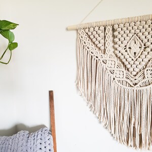 May include: A beige macrame wall hanging with a geometric pattern and fringe. The hanging is attached to a wooden dowel.