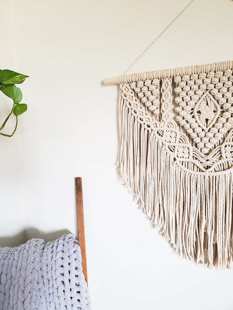 May include: A beige macrame wall hanging with a geometric pattern and fringe. The hanging is attached to a wooden dowel.