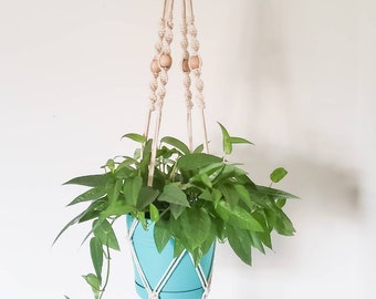 May include: A macrame plant hanger with wooden beads, holding a teal pot with a green plant. The hanger is made of natural cotton rope and has a tassel at the bottom.