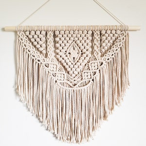 May include: A beige macrame wall hanging with a geometric pattern and fringe. The hanging is attached to a wooden dowel rod.