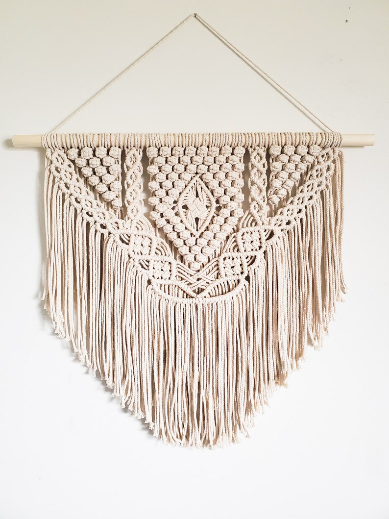 May include: A beige macrame wall hanging with a geometric pattern and fringe. The hanging is attached to a wooden dowel rod.