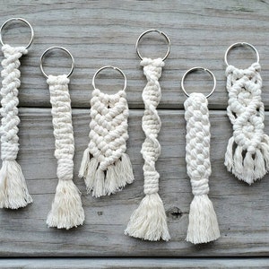 May include: Six white macrame keychains with different patterns and designs. The keychains are made with white cord and have a silver metal ring at the top.