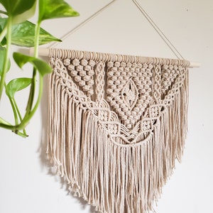 May include: A beige macrame wall hanging with a geometric pattern and fringe. The hanging is attached to a wooden dowel rod.
