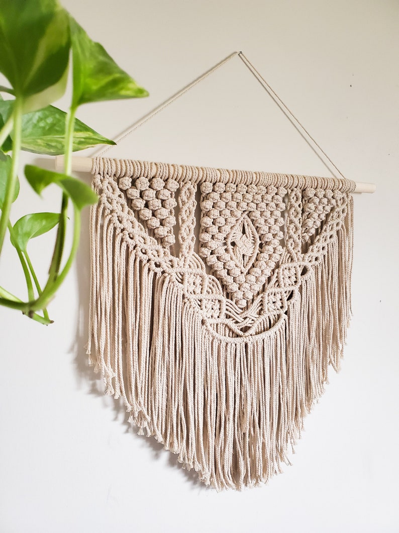 May include: A beige macrame wall hanging with a geometric pattern and fringe. The hanging is attached to a wooden dowel rod.