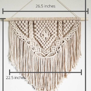 May include: A large, off-white macrame wall hanging with a geometric design. The hanging is 26.5 inches wide, 22.5 inches tall, and 33 inches long. It is made with natural cotton cord and has a wooden dowel at the top.