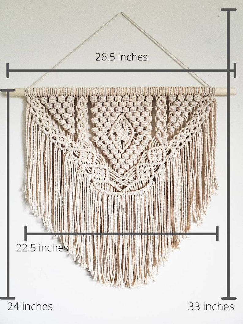 May include: A large, off-white macrame wall hanging with a geometric design. The hanging is 26.5 inches wide, 22.5 inches tall, and 33 inches long. It is made with natural cotton cord and has a wooden dowel at the top.