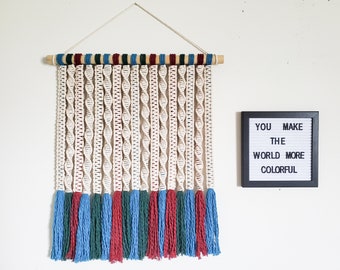 Pode incluir: Uma tapeçaria de macramê branca com franjas vermelhas, azuis e verdes. A tapeçaria está presa a uma haste de madeira. Um quadro de letras preto com o texto "YOU MAKE THE WORLD MORE COLORFUL" está na parede à direita da macramê.