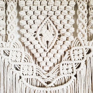 May include: A white macrame wall hanging with a geometric design and fringe. The hanging is made with thick cotton cord and has a wooden dowel at the top.