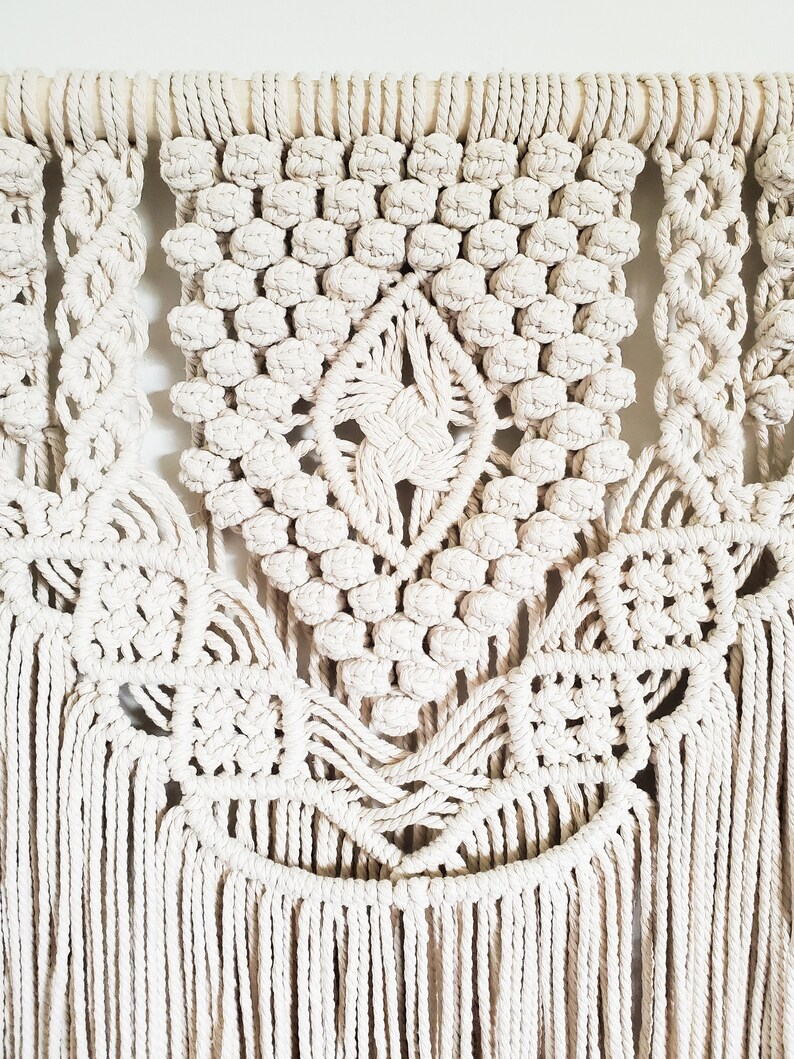 May include: A white macrame wall hanging with a geometric design and fringe. The hanging is made with thick cotton cord and has a wooden dowel at the top.