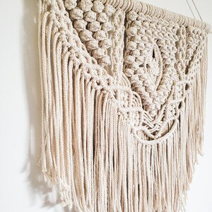 May include: A large, off-white macrame wall hanging with a geometric pattern and long fringe. The hanging is made with natural cotton cord and is attached to a wooden dowel.