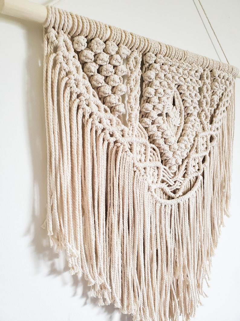 May include: A large, off-white macrame wall hanging with a geometric pattern and long fringe. The hanging is made with natural cotton cord and is attached to a wooden dowel.