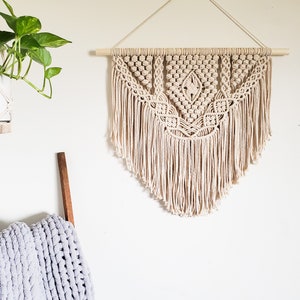 May include: A beige macrame wall hanging with a geometric pattern and fringe. The hanging is attached to a wooden dowel rod.