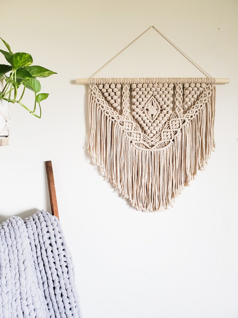 May include: A beige macrame wall hanging with a geometric pattern and fringe. The hanging is attached to a wooden dowel rod.