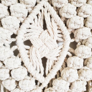 May include: Close-up of a white macrame wall hanging with a diamond-shaped center and a textured, knotted design.