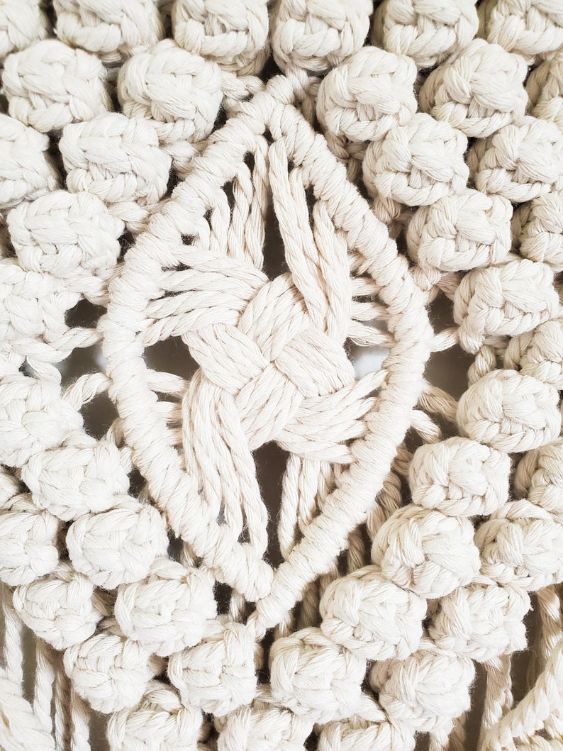 May include: Close-up of a white macrame wall hanging with a diamond-shaped center and a textured, knotted design.