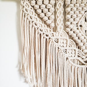 May include: A beige macrame wall hanging with a geometric pattern and fringe. The hanging is made with natural cotton cord and is attached to a wooden dowel.
