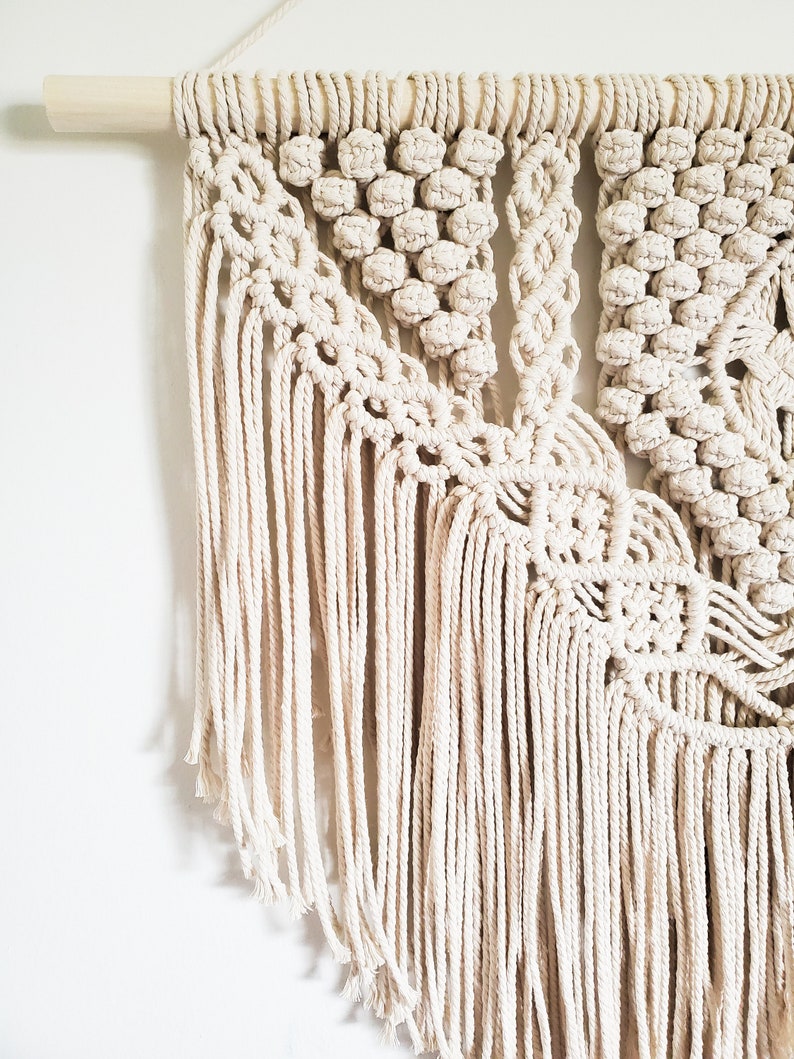 May include: A beige macrame wall hanging with a geometric pattern and fringe. The hanging is made with natural cotton cord and is attached to a wooden dowel.