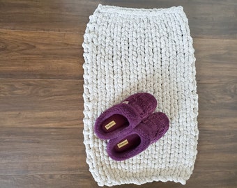 May include: A pair of purple fuzzy slippers on a gray knitted rug. The rug is made of thick, chunky yarn and has a textured surface.