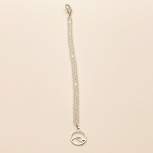 May include: A beaded bracelet featuring a single strand of translucent, iridescent glass beads. The bracelet is finished with a silver-toned metal lobster claw clasp at one end and a circular silver-toned metal charm depicting a wave at the other end.