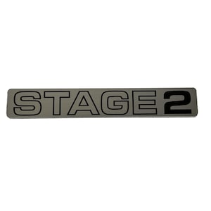May include: A silver metal plaque with black lettering that reads "STAGE 2".