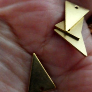 Op de afbeelding: Drie gouden driehoekige metalen bedels met gaten aan de bovenkant. De bedels liggen op een hand.
