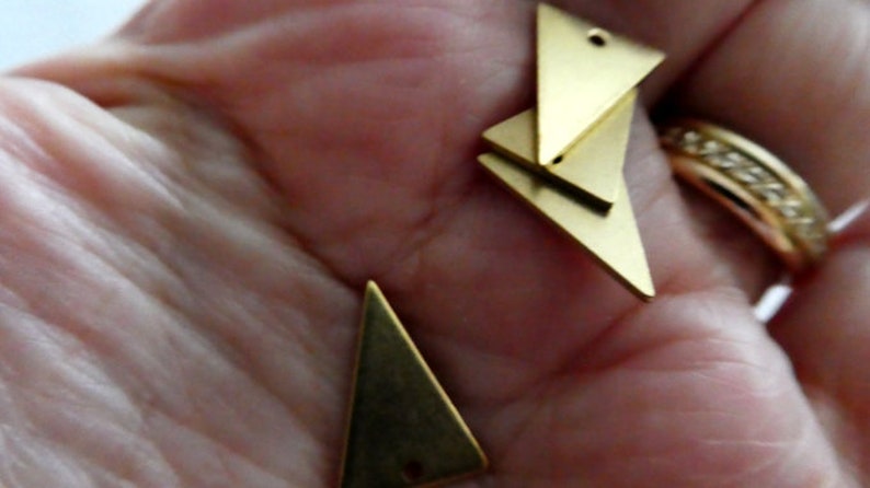 Op de afbeelding: Drie gouden driehoekige metalen bedels met gaten aan de bovenkant. De bedels liggen op een hand.