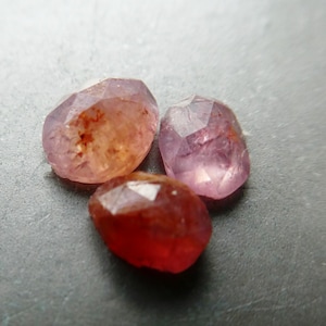 May include: Three faceted gemstones in shades of red, pink, and orange are displayed. The gems have a slightly irregular shape and a polished, reflective surface. The stones are arranged closely together, showcasing their color variations.