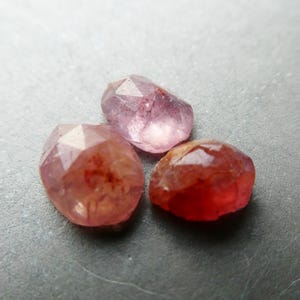 May include: Three faceted gemstones in shades of pink and red. The gems have irregular shapes and a polished, reflective surface. The stones are arranged on a gray surface, showcasing their color variations.