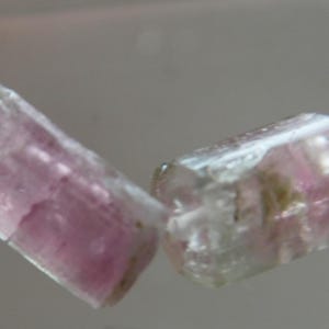 May include: Two raw, pink tourmaline gemstones are strung on a thin, clear cord. The gemstones are rectangular with a translucent appearance, showcasing a blend of pink and clear hues. The stones are rough-cut, displaying natural textures.