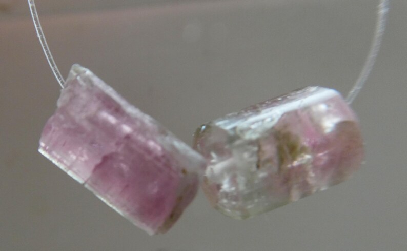 May include: Two raw, pink tourmaline gemstones are strung on a thin, clear cord. The gemstones are rectangular with a translucent appearance, showcasing a blend of pink and clear hues. The stones are rough-cut, displaying natural textures.
