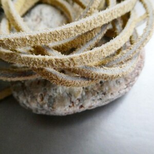 May include: Close-up of a coiled, light tan leather cord resting on a textured, light-colored stone. The cord appears to be made of multiple strands, with a slightly fuzzy texture. The stone has a rough, porous surface.