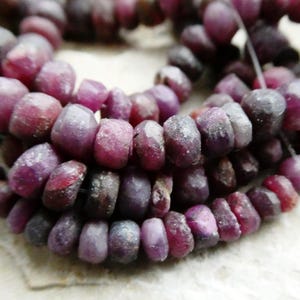 May include: Strands of natural ruby gemstone beads. The beads are a deep reddish-purple color, with variations in tone. The beads are irregularly shaped and have a faceted appearance. The beads are strung together on a thin, clear string.
