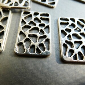 May include: Close-up of several rectangular silver-toned metal charms. Each charm has an open, organic pattern of irregular shapes within its frame. The charms are scattered on a grey surface, with some slightly out of focus.