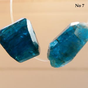 May include: Two faceted, teal-colored gemstone beads are strung on a thin, clear cord. The gemstones have a crystalline structure and are irregularly shaped. The text "No 7" is in the upper right corner.