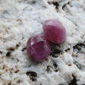 May include: Two faceted, oval-shaped, deep red gemstones. The gems are set against a rough, light-colored stone surface. The gems have a polished, reflective surface, and the facets catch the light, creating a sparkling effect. The overall theme is gemstones.