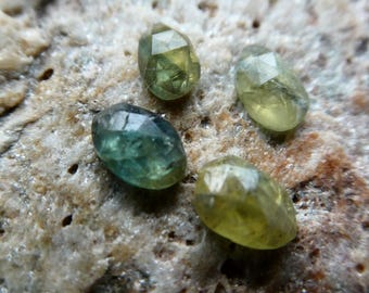 May include: Four small, faceted, oval-shaped gemstones in shades of olive green and teal. Each stone features a polished, multi-faceted surface that reflects light, showcasing a translucent quality and distinct color variations across the set.
