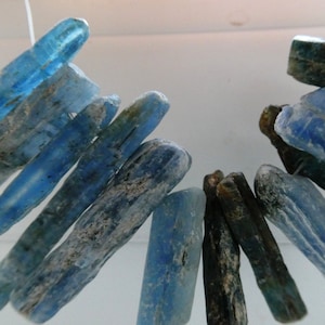 May include: A collection of blue and green kyanite crystal points, some with a white cloudy appearance, arranged in a semi-circle shape.